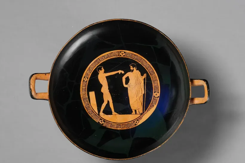 Fotografia de uma taça grega de barro, preta por fora, com duas alças; no medalhão central, cercado por uma greca em ziguezague, um rapaz nu de perfil estende o braço para um homem barbado de manto longo, que segura um bastão.