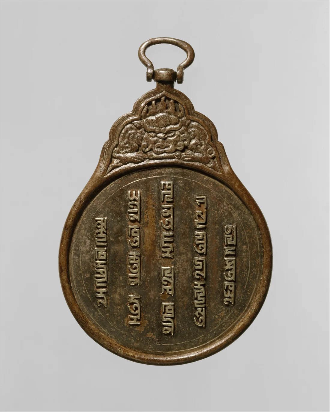 Fotografia de uma placa de ferro escurecido em forma de pera, com uma argola no alto; o topo traz em relevo a cabeça de uma fera de juba, e o disco é coberto por cinco colunas verticais de caracteres em escrita phags-pa, incrustados em prata.