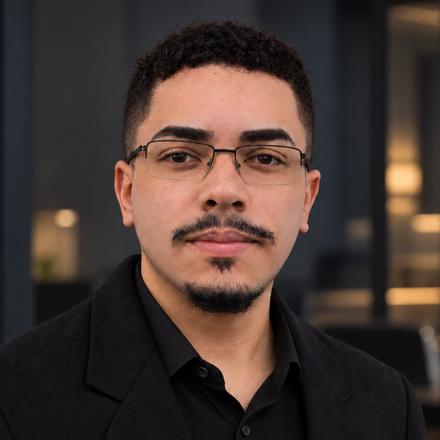 Retrato de Alex Mërović: homem de cabelo curto e cacheado, barba e bigode aparados e óculos de armação fina, de blazer preto sobre camisa preta, em ambiente interno desfocado